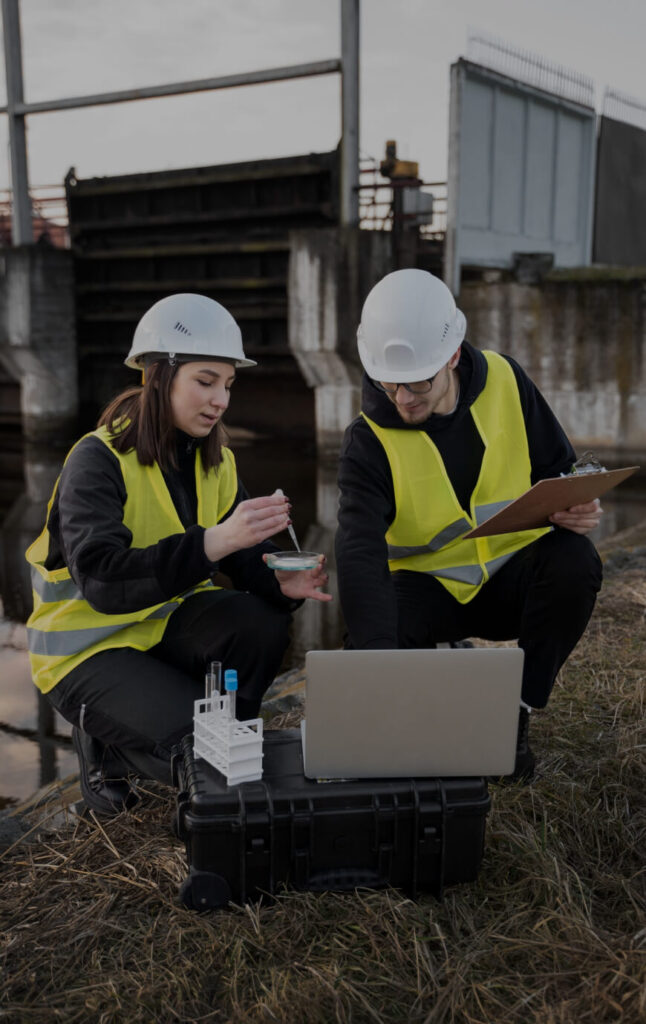 Two restoration professionals in safety gear conducting water damage assessment, analyzing samples, and documenting findings on a laptop, with a waterway and industrial structure in the background.