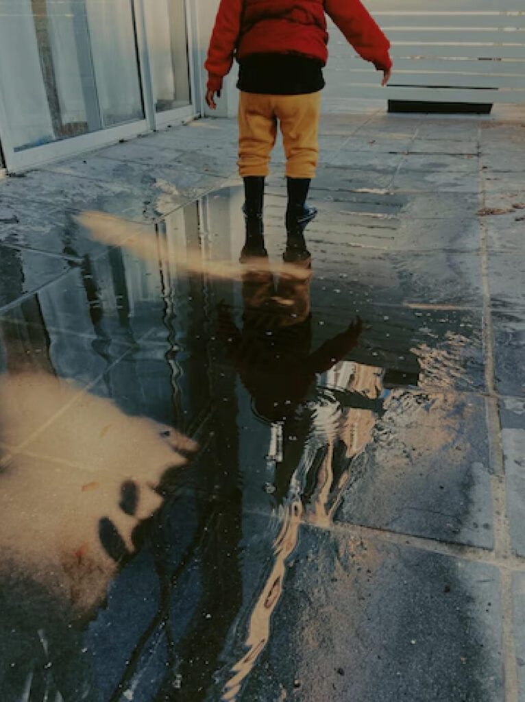 Child in red jacket and yellow pants standing in water puddle on stone surface, reflecting surroundings, illustrating water damage scenario relevant to restoration services.
